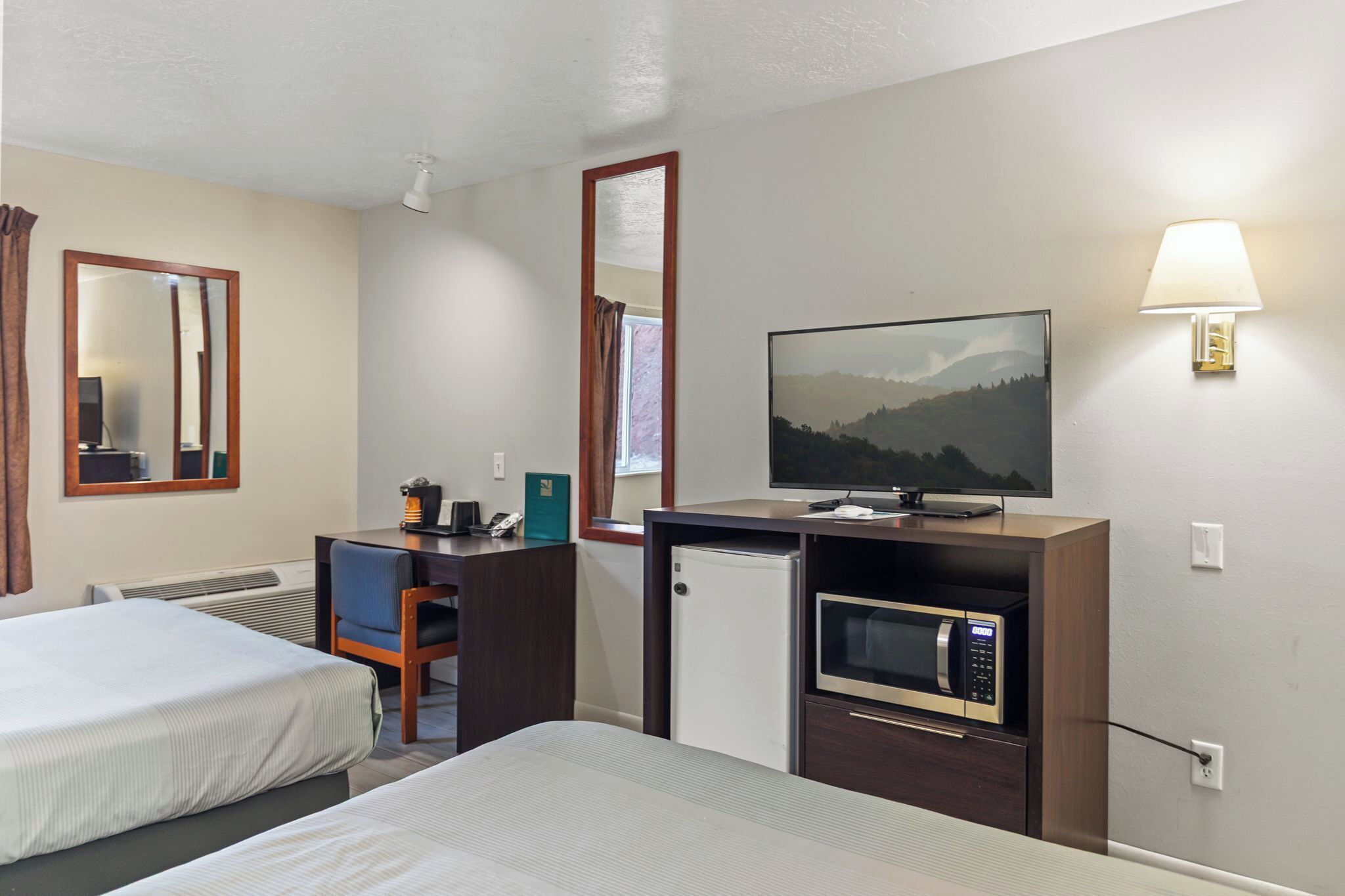 A hotel room with two beds, a desk with a computer, a microwave, a TV, and a wall-mounted lamp.