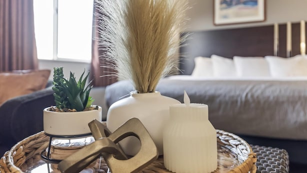 A wicker tray with a plant, a candle, and a metal object on a bed with a headboard and pillows.