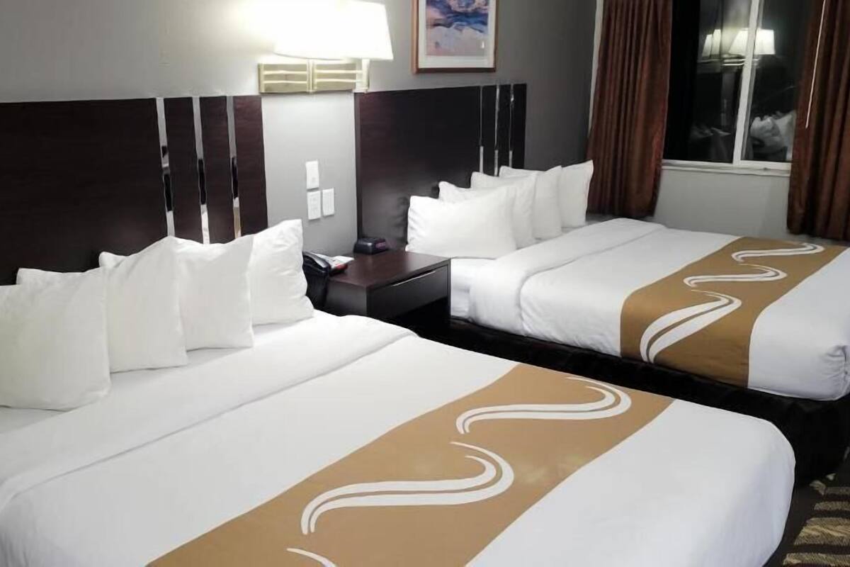 A hotel room with two beds, a nightstand, a lamp, and a window with curtains.