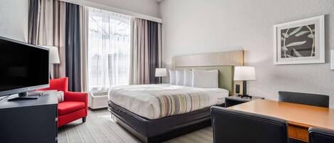 Quarto de hotel com cama, escrivaninha, televisão, uma cadeira vermelha e uma janela grande com cortinas.