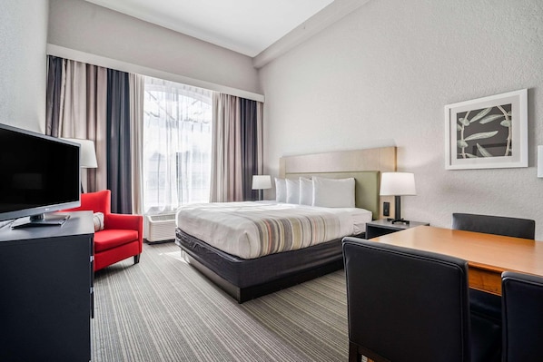 Quarto de hotel com cama, escrivaninha, televisão, uma cadeira vermelha e uma janela grande com cortinas.