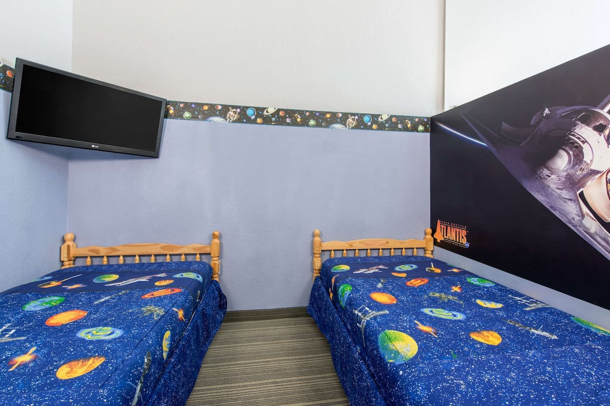 A room with two single beds, a flat-screen TV mounted on the wall, and a large poster of a space shuttle on the right wall.