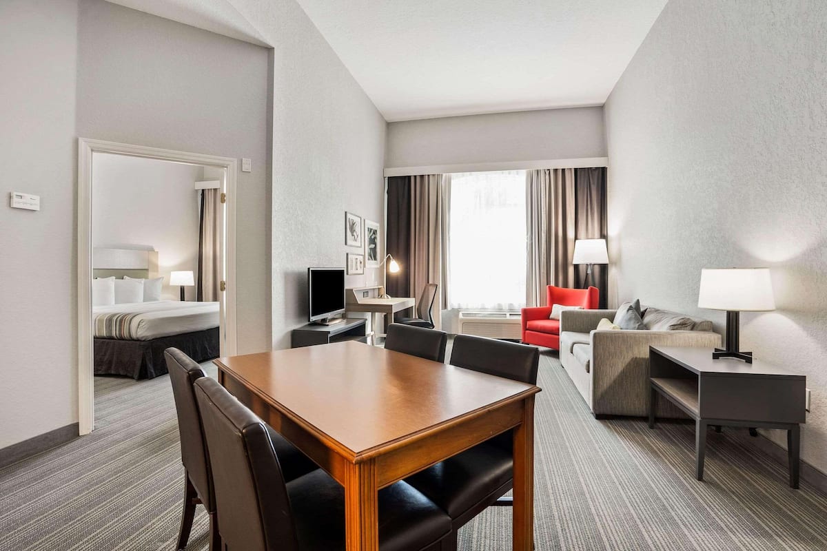 A hotel room with a dining area, a living area with a sofa and armchairs, a TV, and a bed.