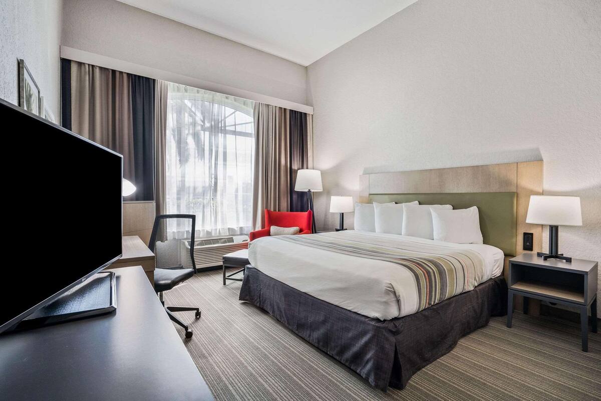 A hotel room with a large bed, a desk with a flat-screen TV, a chair, two bedside tables with lamps, and a window with curtains.