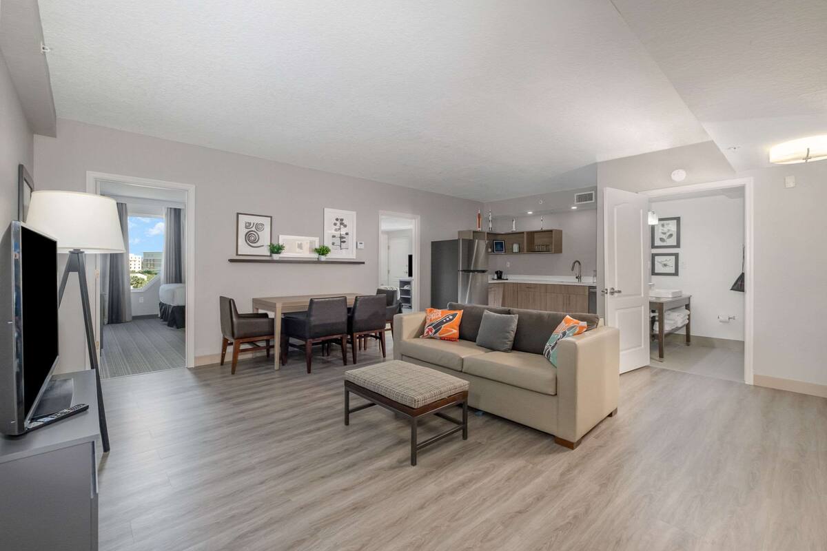 A modern living room with a sofa, a coffee table, and a TV. There is a dining area with chairs and a table, and a kitchen with cabinets and a refrigerator.