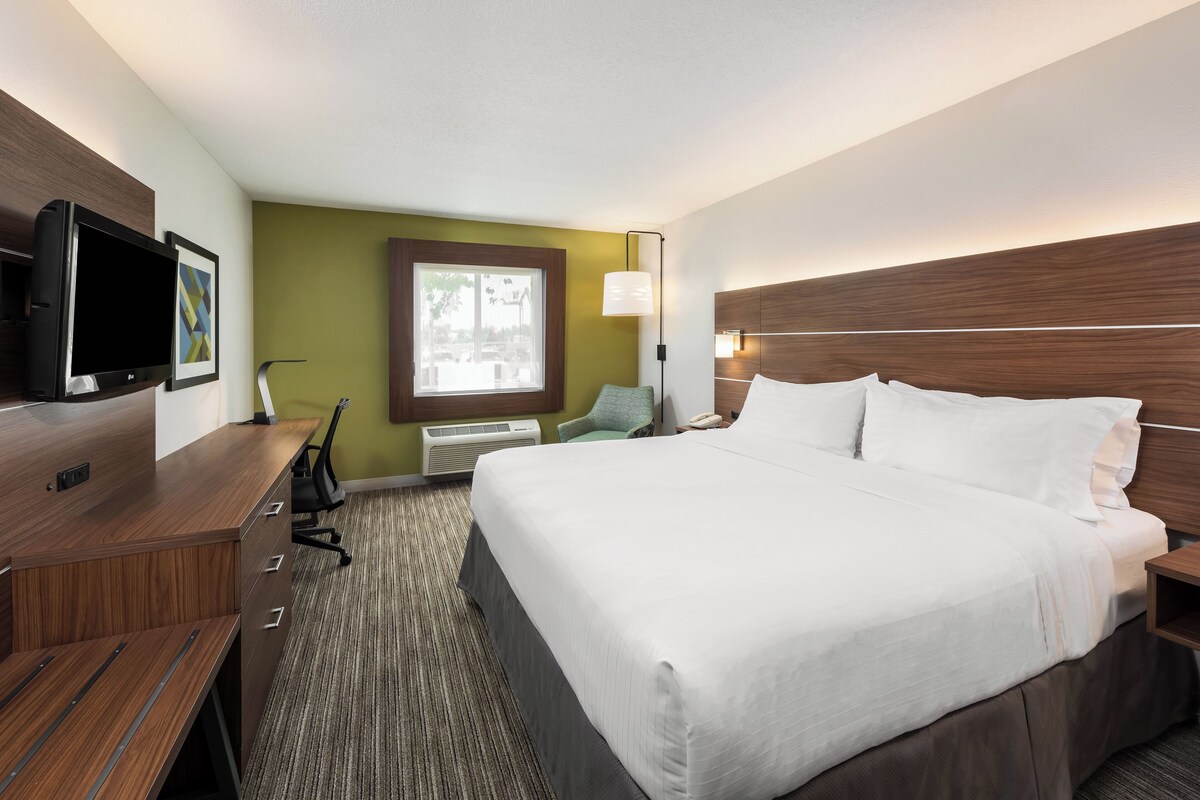 A hotel room with a large bed, a desk with a flat-screen TV, a chair, a window with a view, and a wall-mounted lamp.