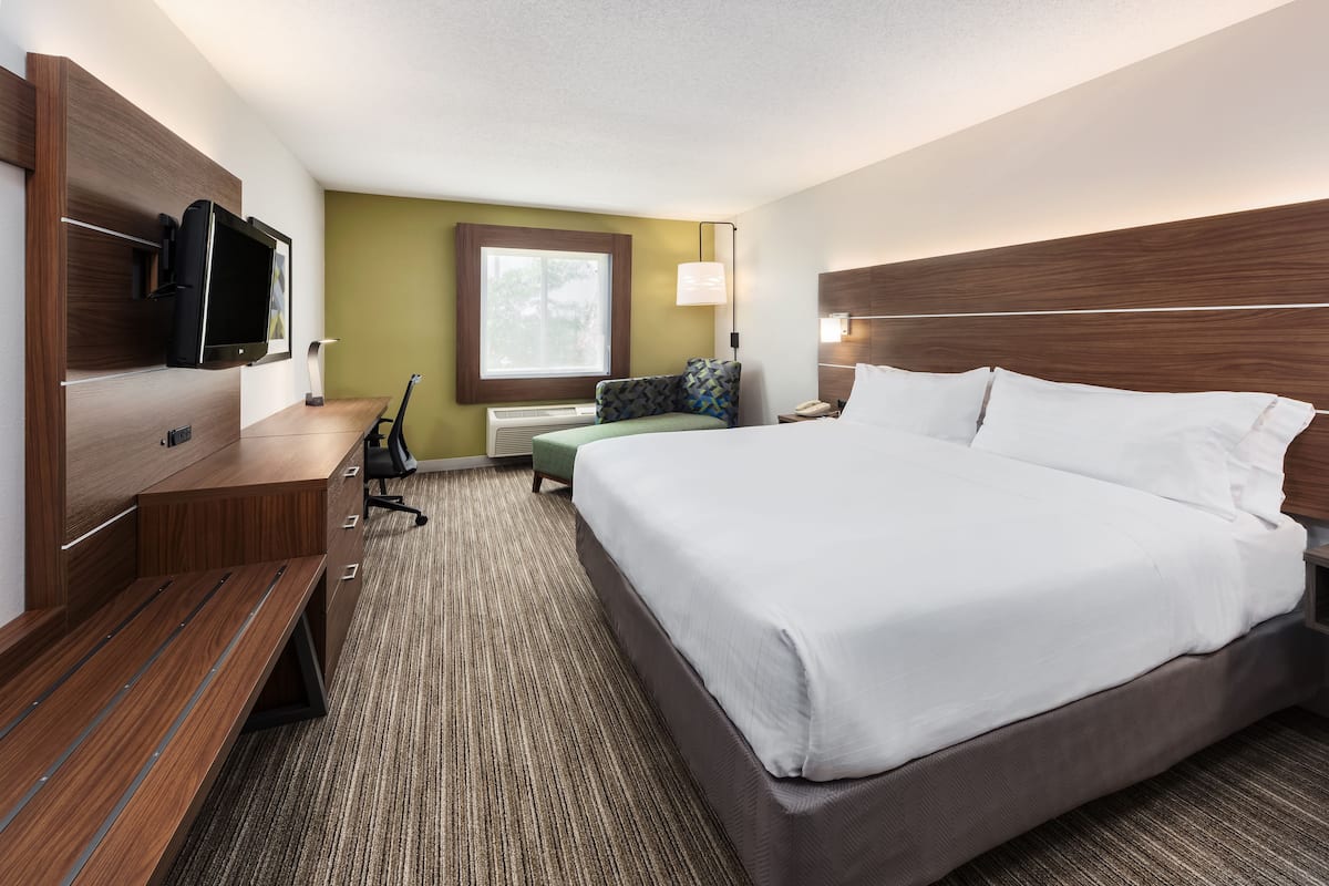 A hotel room with a large bed, a desk with a flat-screen TV, a chair, and a window with a view of trees.