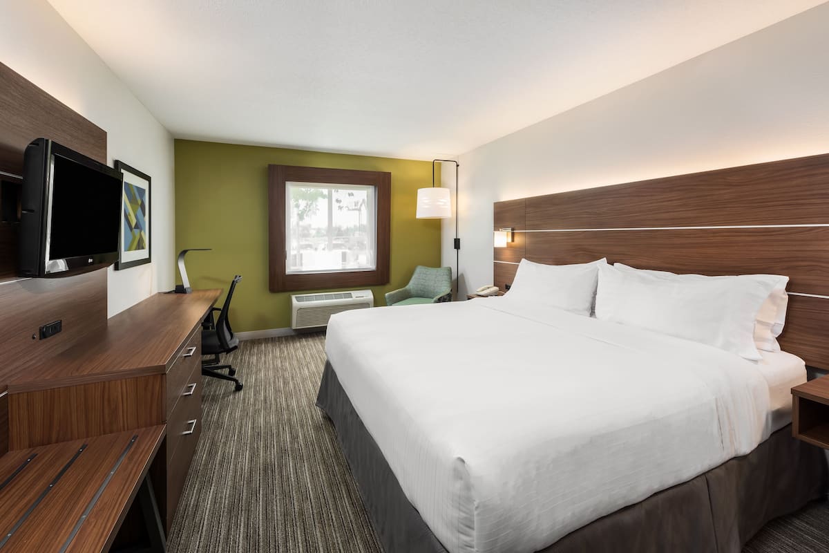 A hotel room with a large bed, a desk with a flat-screen TV, a chair, a window with a view, and a wall-mounted lamp.