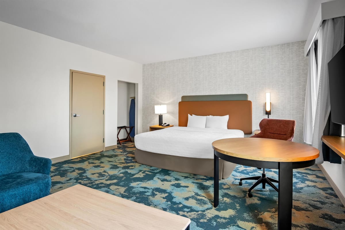 Habitación de hotel con cama, una mesa redonda, un sillón azul, un escritorio de madera y un televisor sobre un mueble.