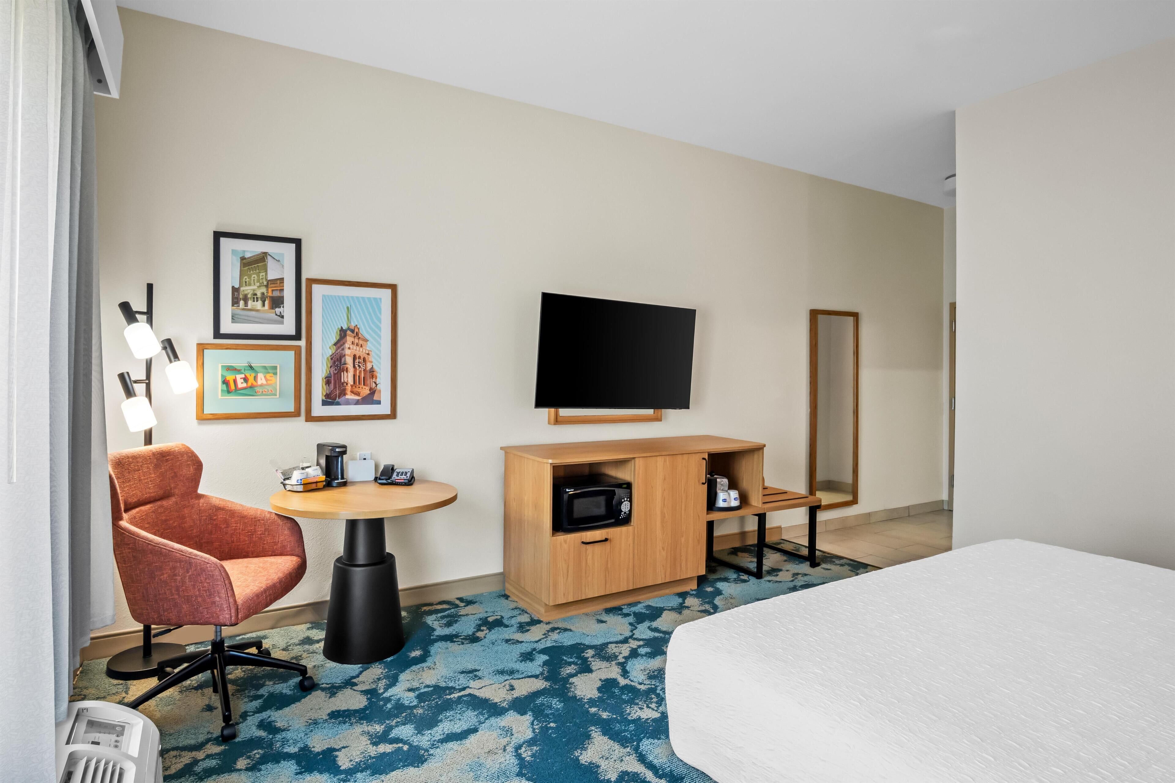 A hotel room with a bed, a desk with a computer, a chair, a microwave, a television, and framed pictures on the wall.