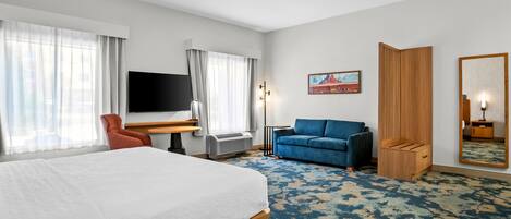 A hotel room with a large bed, a desk with a flat-screen TV, a blue sofa, a wooden wardrobe, and a large mirror.