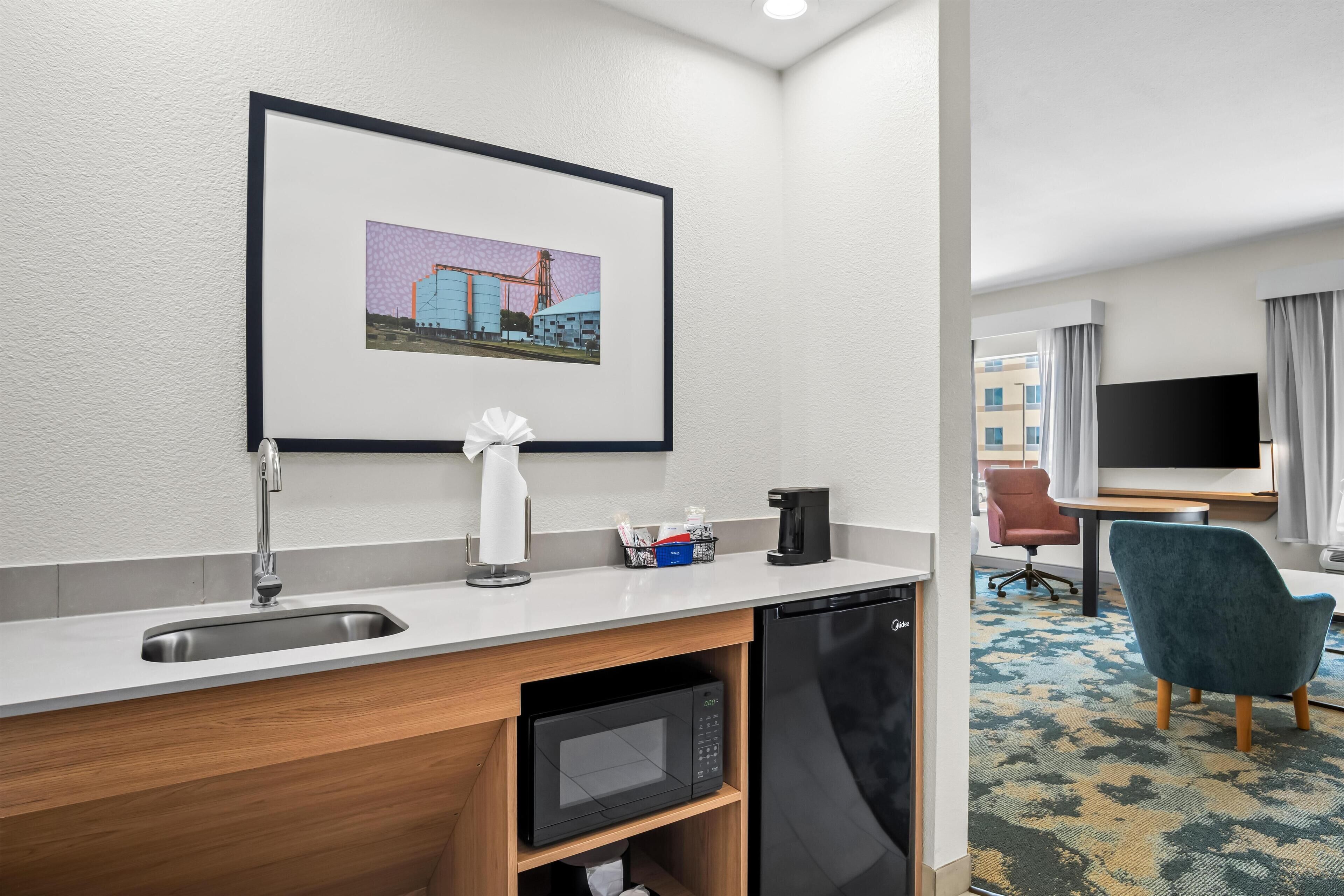 Una habitación de hotel con kitchenette, un cuadro en la pared, un escritorio con silla y un televisor.