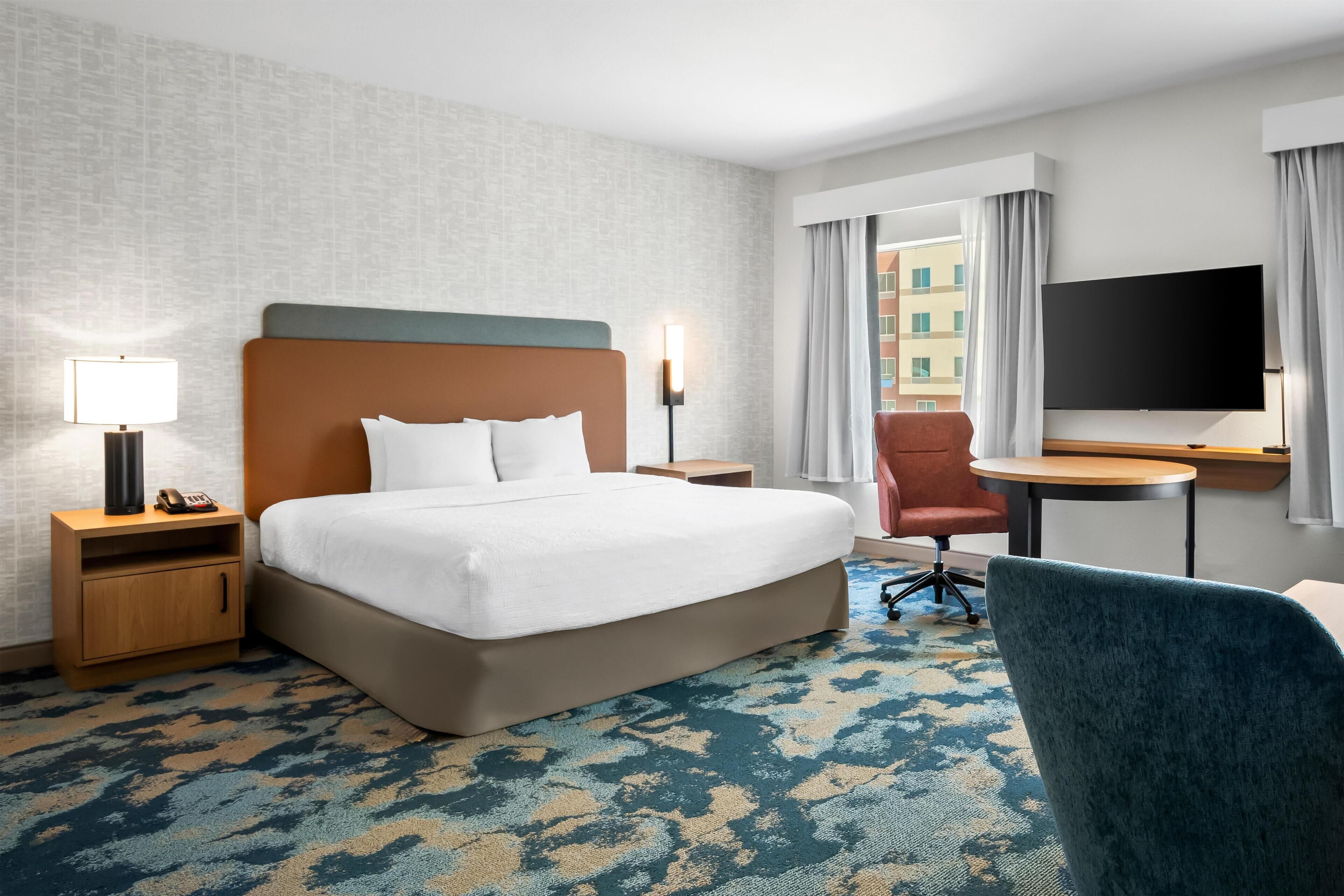 A hotel room with a large bed, a desk with a chair, a TV, and a window with curtains.