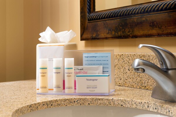 Tocador de baño con artículos de aseo personal, incluyendo productos Neutrogena, pañuelos de papel y un cartel sobre objetos olvidados.