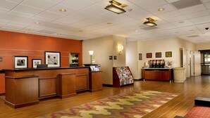Un espace d’accueil d’hôtel avec un comptoir de réception en bois, un panneau d’accueil, des tableaux encadrés, un coin café et une moquette à motifs.