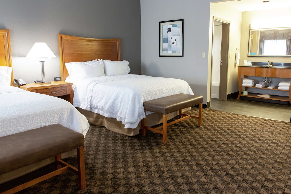 A hotel room with two beds, a bench, a bedside table with a lamp, a framed picture on the wall, and a bathroom with a sink and towel rack.