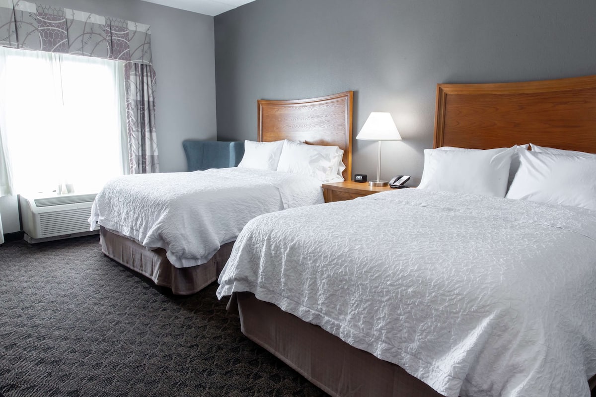 A hotel room with two beds, a nightstand with a lamp, and a window with curtains.