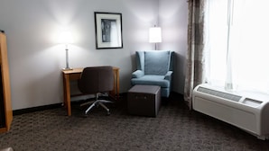 A hotel room with a desk, chair, ottoman, air conditioning unit, and a framed picture on the wall.