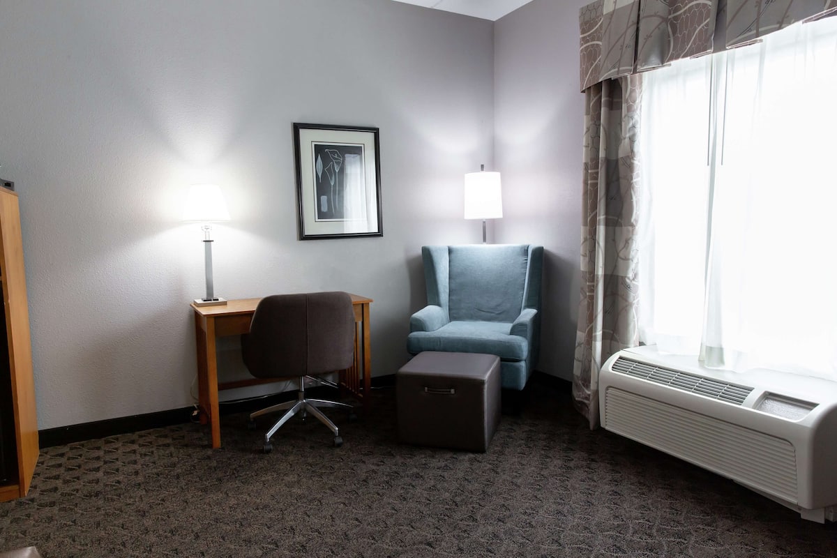 A hotel room with a desk, chair, ottoman, air conditioning unit, and a framed picture on the wall.