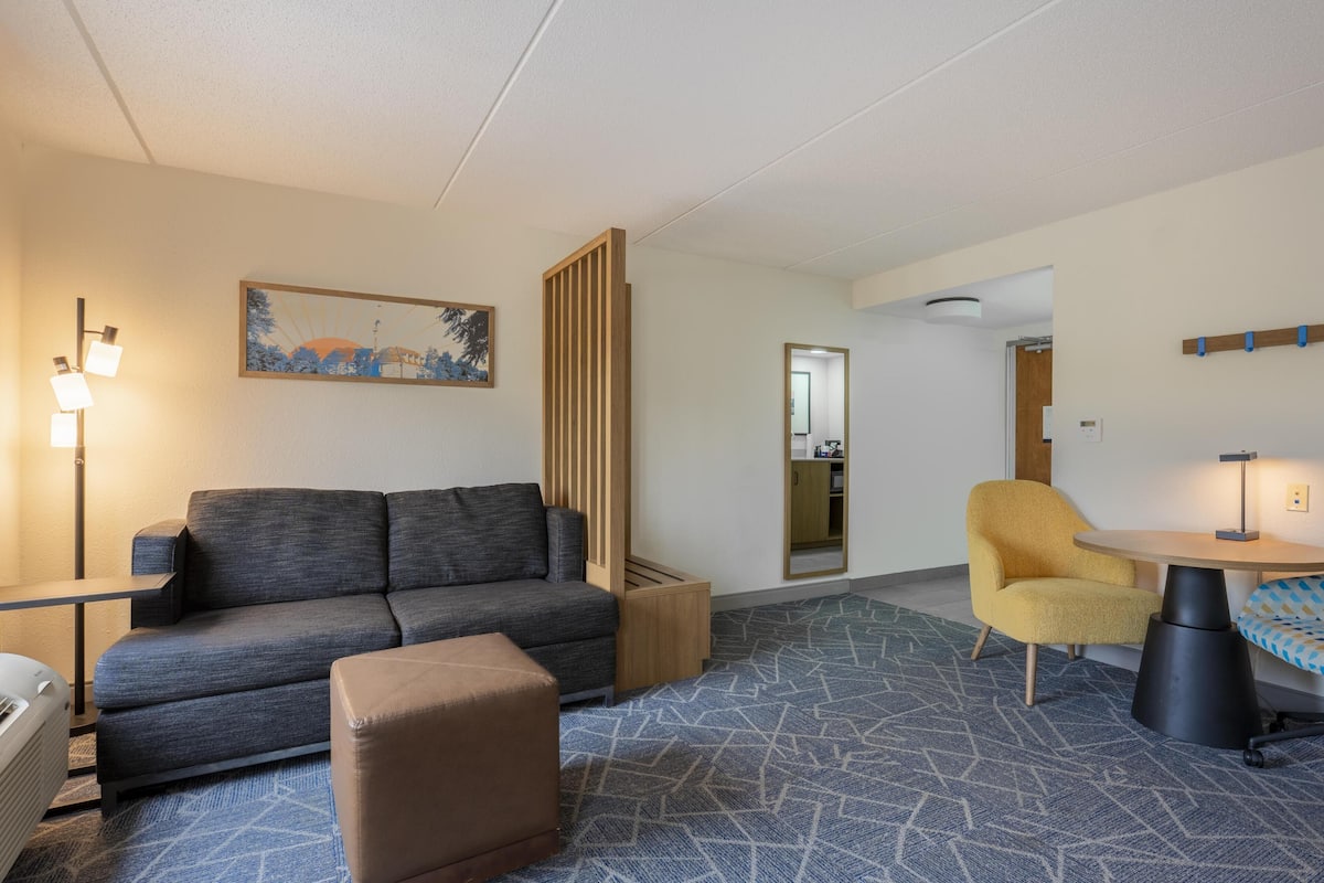 Een moderne hotelkamer met een grijze bank, een klein voetenbankje, een gele fauteuil, een ronde tafel en een kamerbreed tapijt met een patroon.