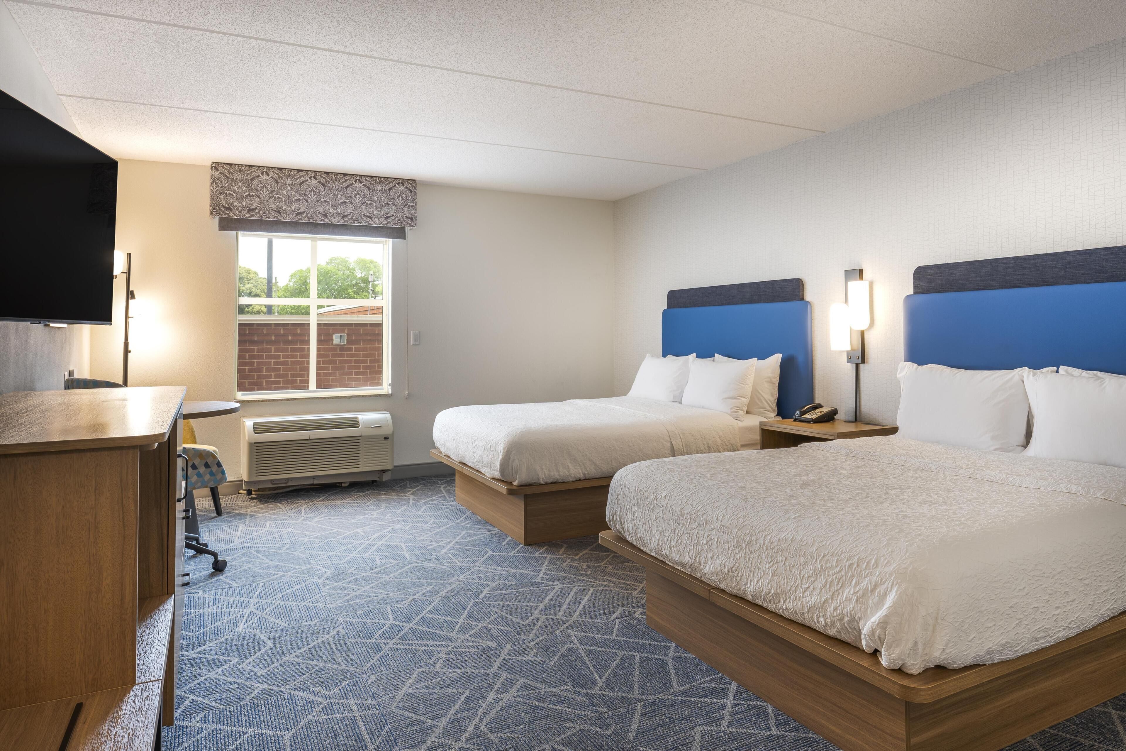 Quarto de hotel com duas camas, uma escrivaninha, uma cadeira, uma TV e uma janela com vista para o exterior.