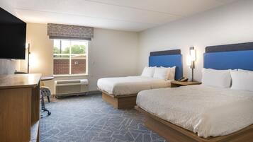 Quarto de hotel com duas camas, uma escrivaninha, uma cadeira, uma TV e uma janela com vista para o exterior.
