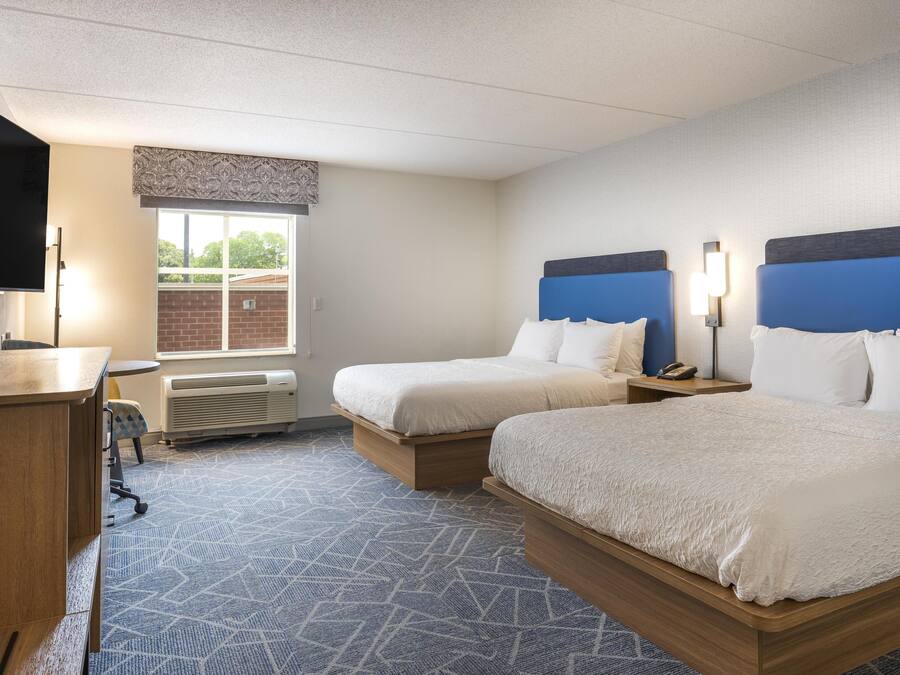Quarto de hotel com duas camas, uma escrivaninha, uma cadeira, uma TV e uma janela com vista para o exterior.