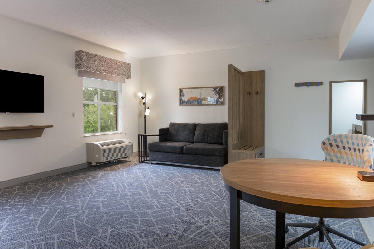 Een moderne woonkamer met een bank, een ronde tafel, een aan de muur bevestigde televisie en een raam met uitzicht op bomen.
