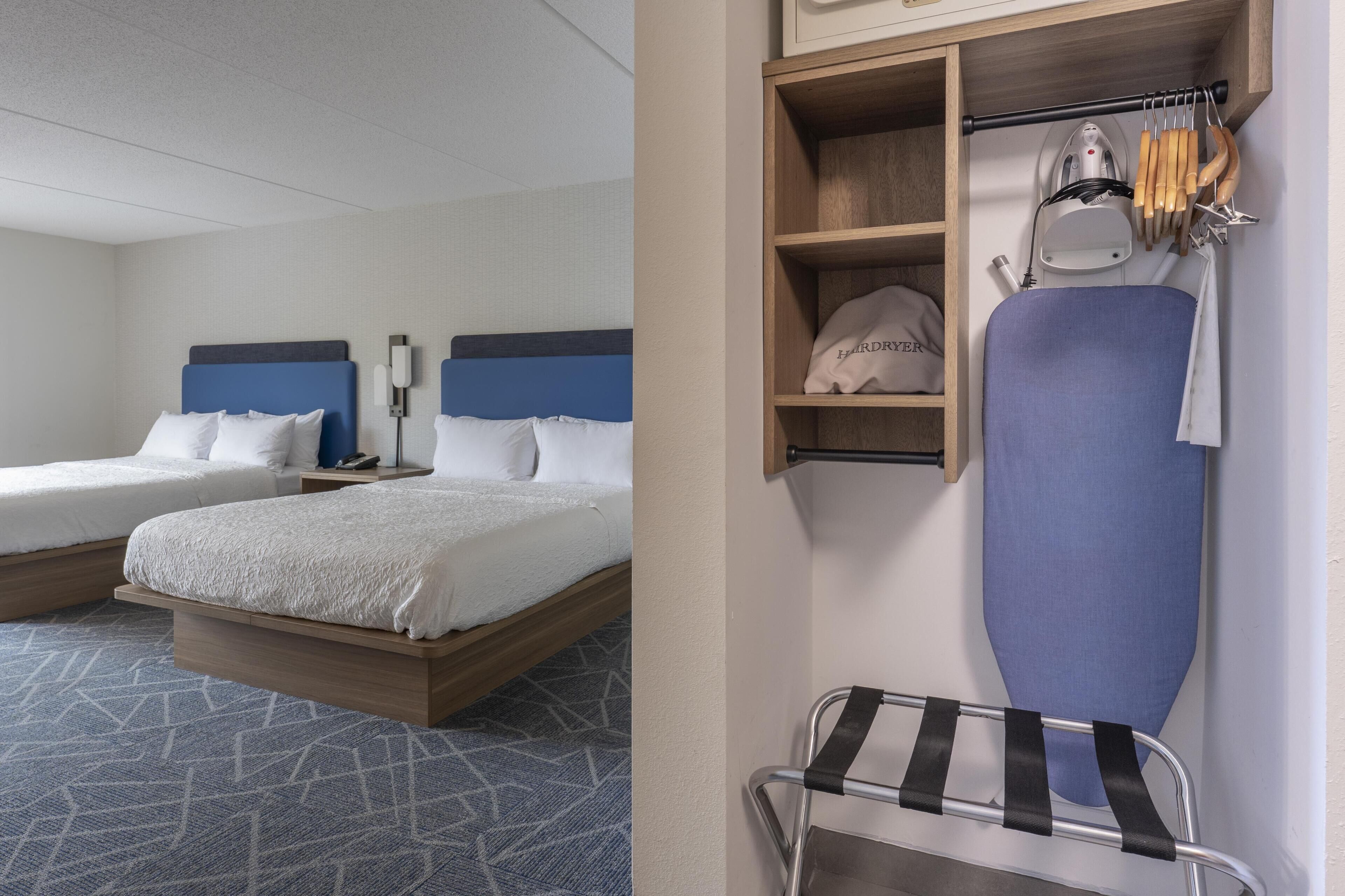 A hotel room with two beds, a wardrobe with hanging clothes and an ironing board.