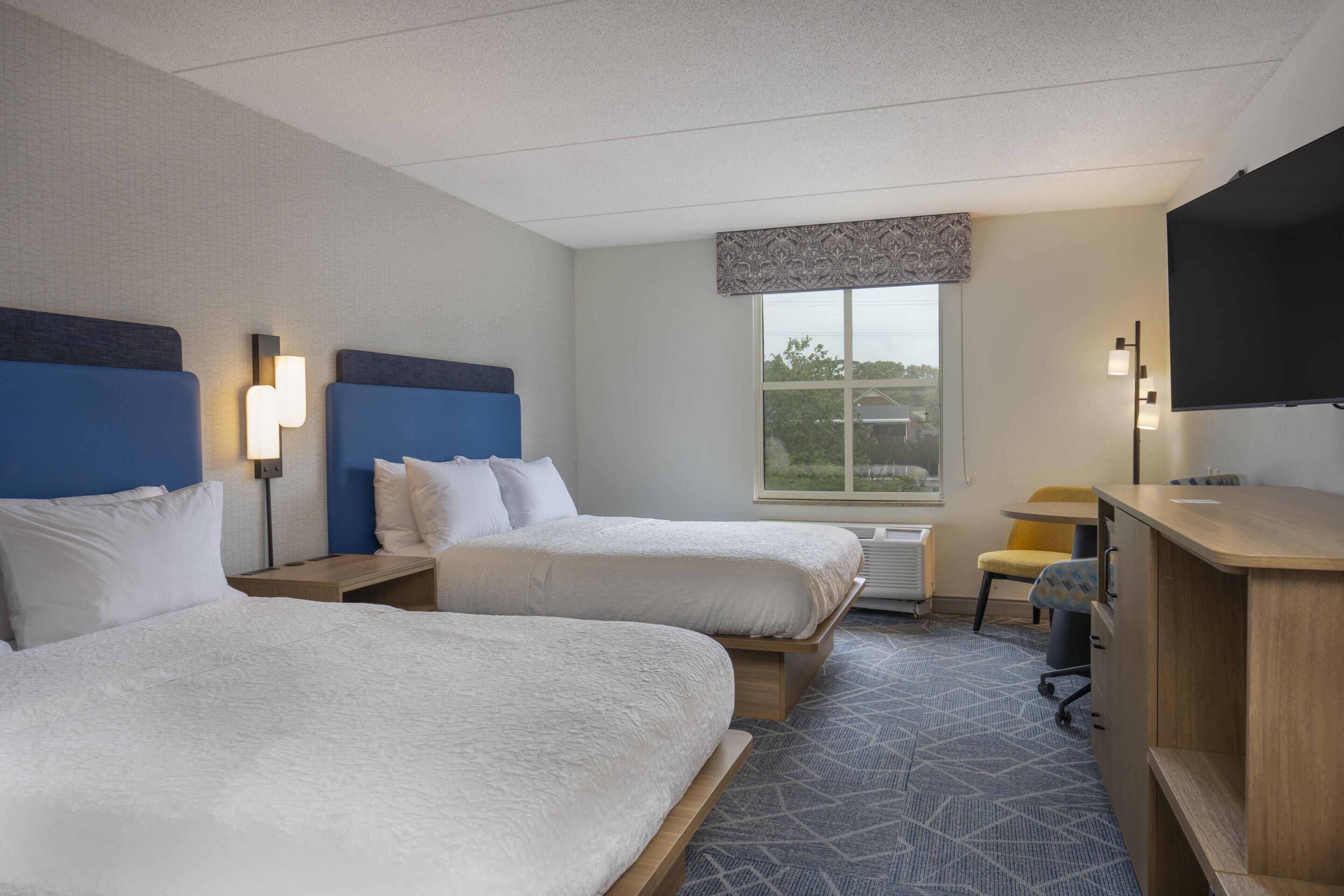 A hotel room with two beds, a desk, a chair, a TV, and a window with a view.