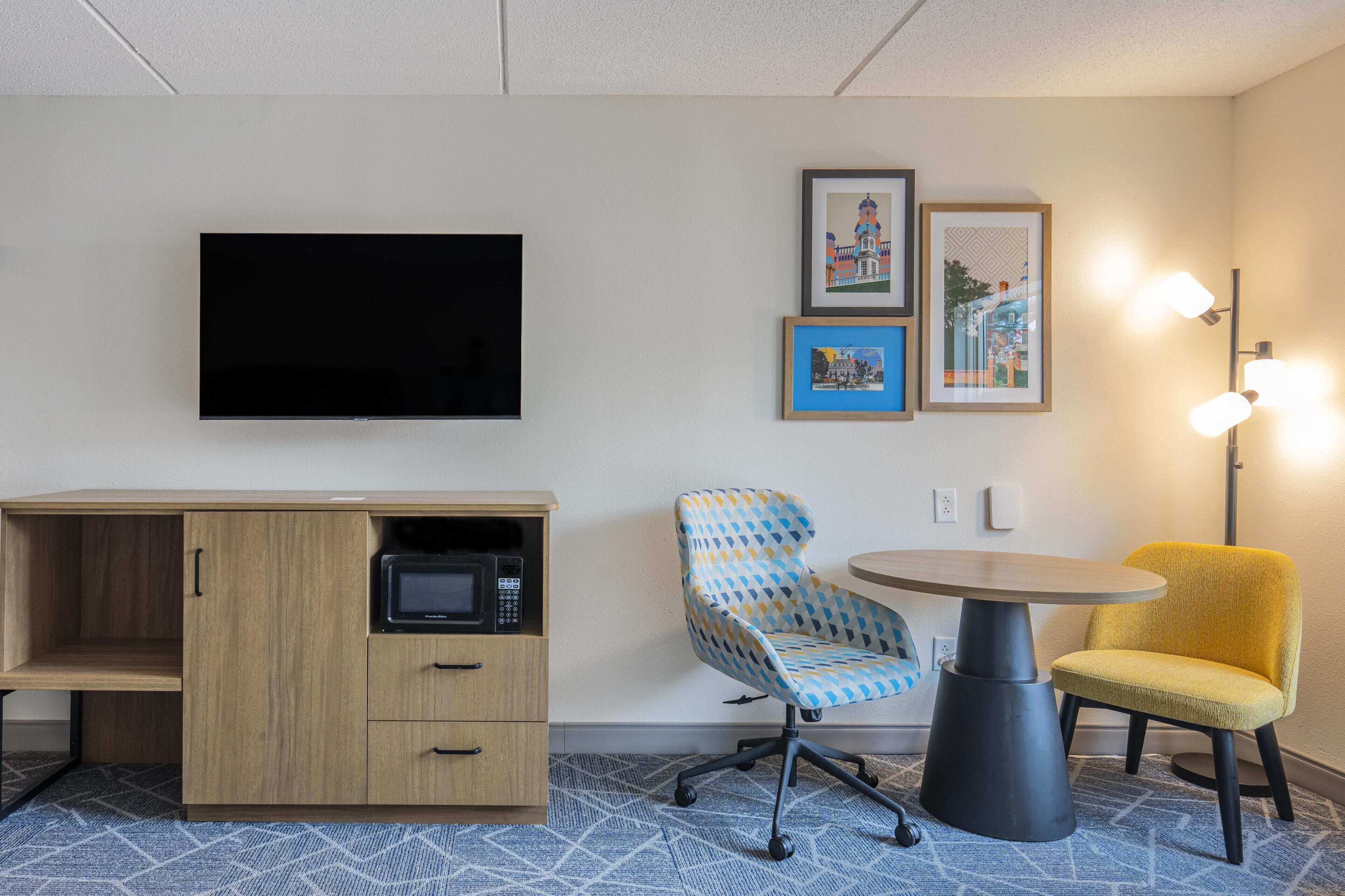 A hotel room with a flat-screen TV, a wooden cabinet with a microwave, a patterned chair, a yellow chair, and a round table.
