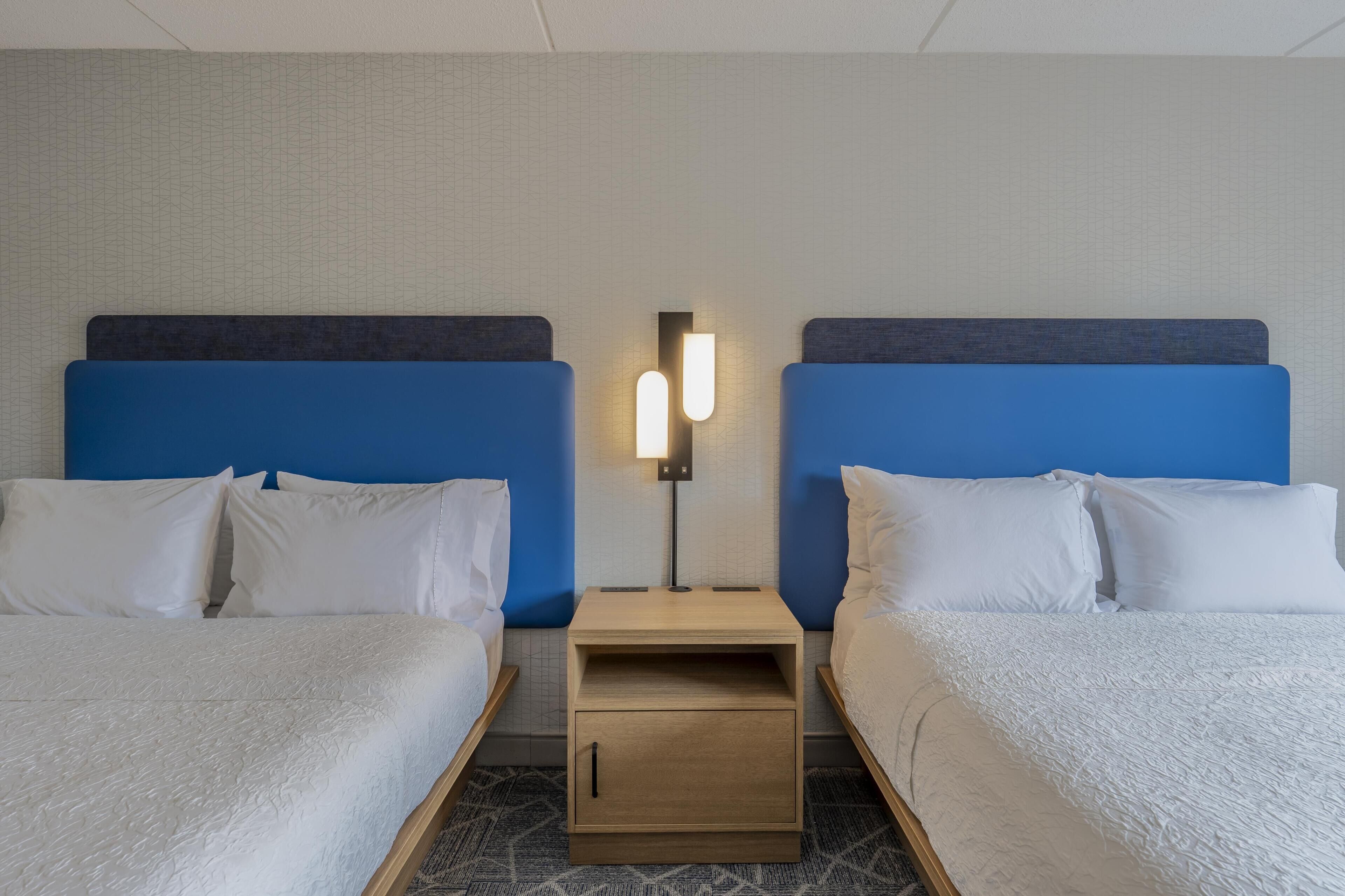 Habitación de hotel con dos camas, una mesita de noche con lámpara y una alfombra estampada.