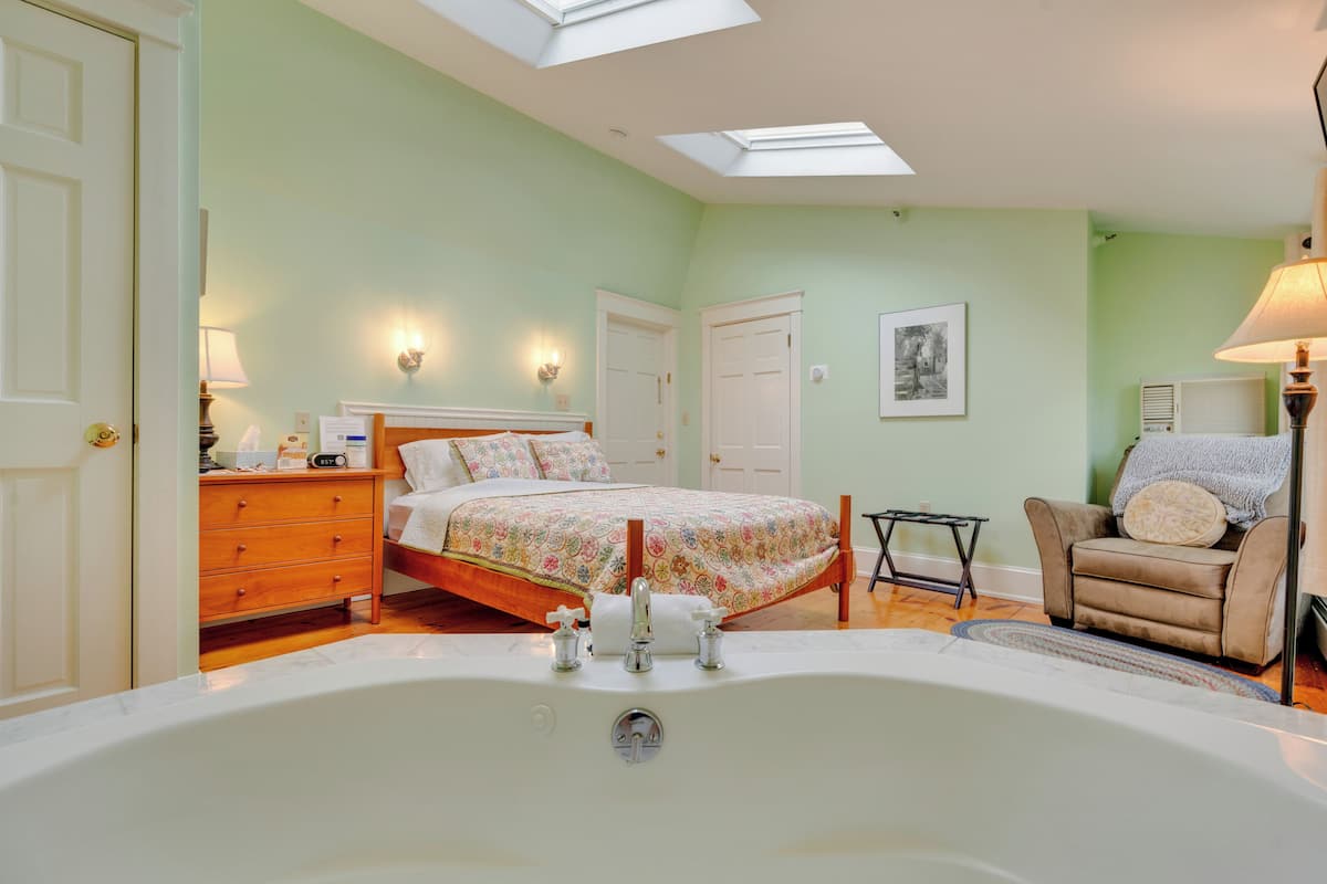 A bedroom with a large bathtub, a bed with floral bedding, a chair, and a small table.