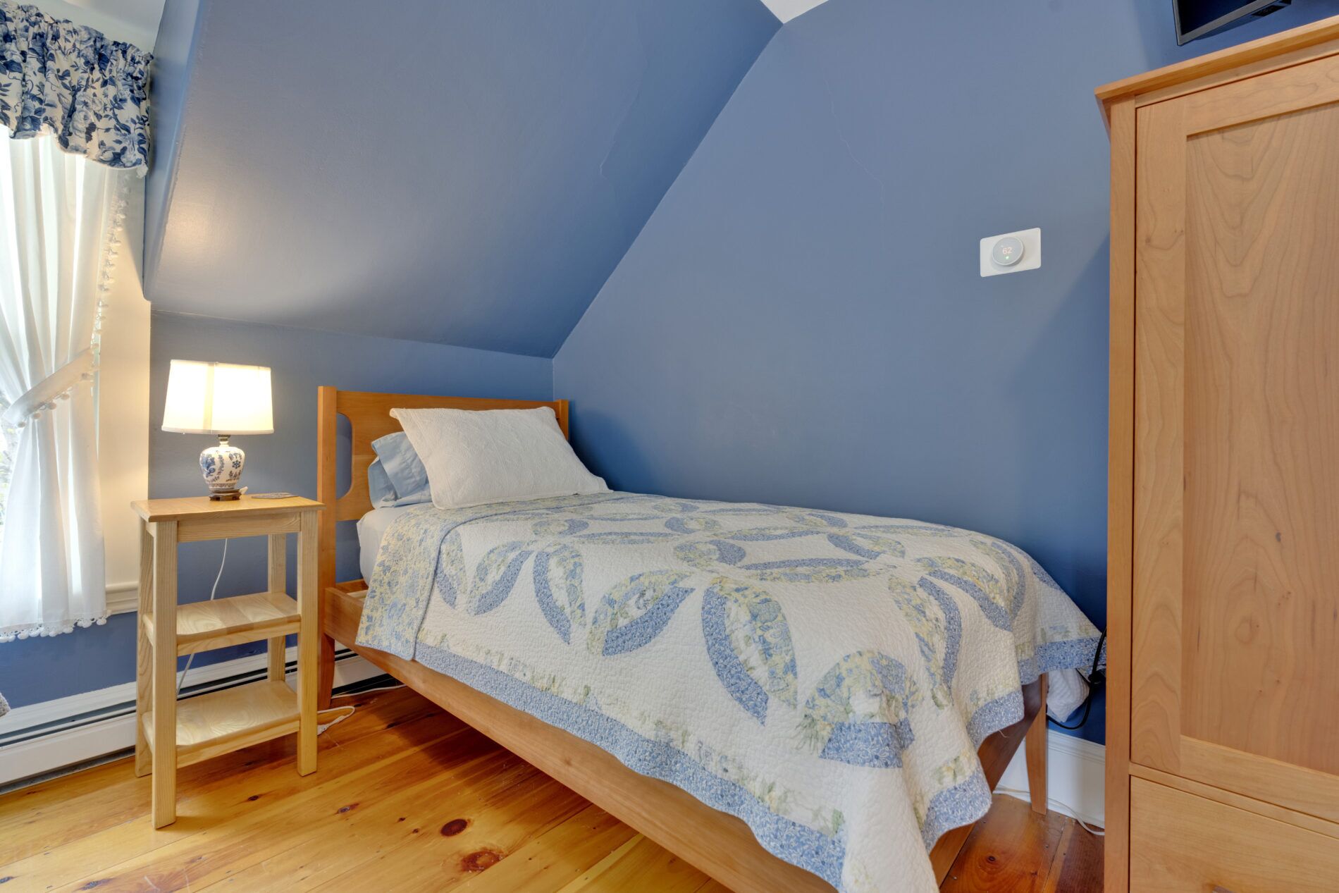 Un dormitorio con una cama individual, una mesita de noche con lámpara, un armario de madera y una ventana con cortinas.