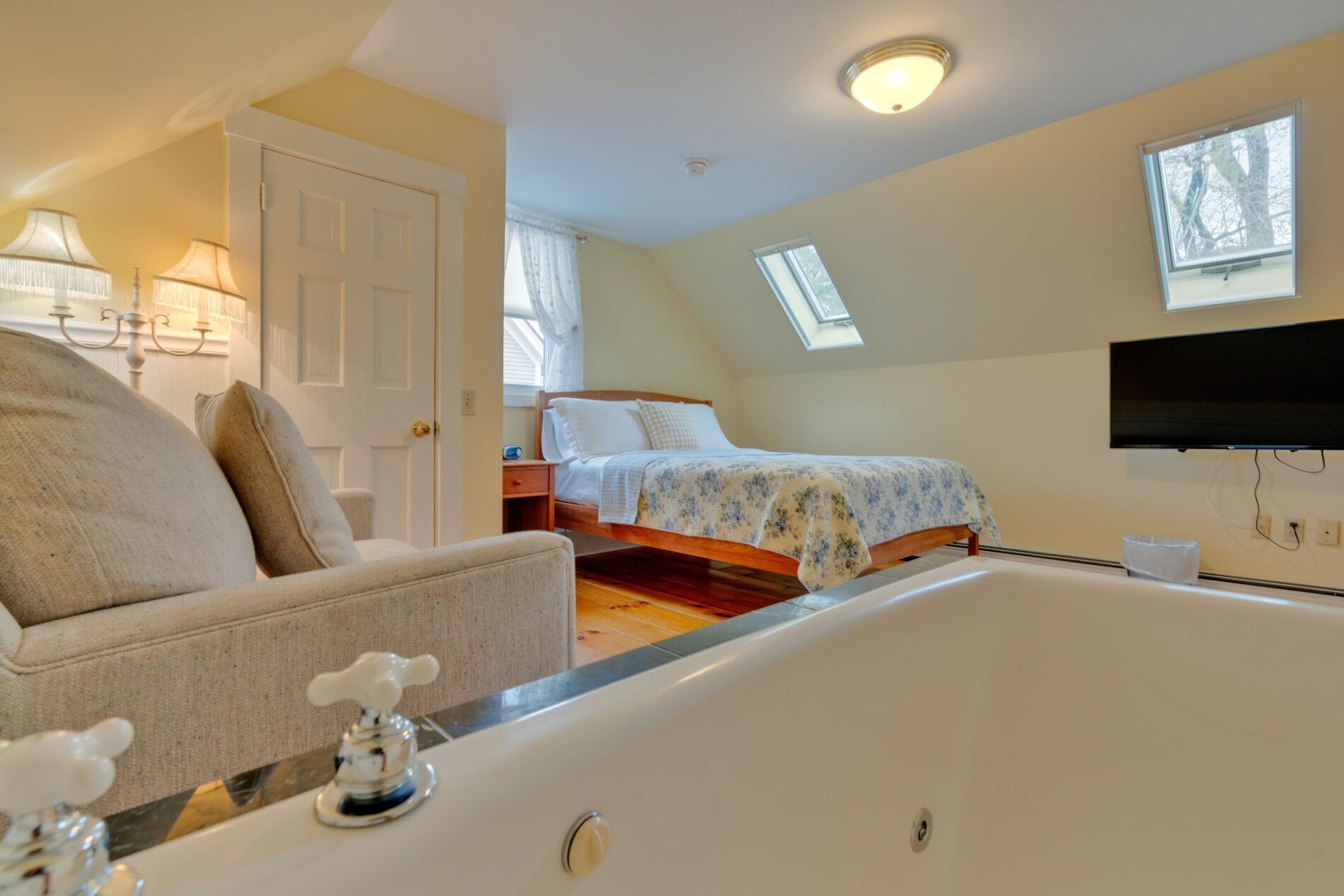 A bedroom with a large bathtub, a bed with floral bedding, a TV mounted on the wall, and a skylight.