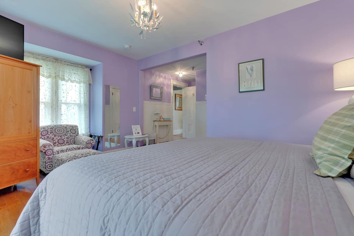 A bedroom with a large bed, a wooden wardrobe, a patterned armchair, a small table, and a mirror.
