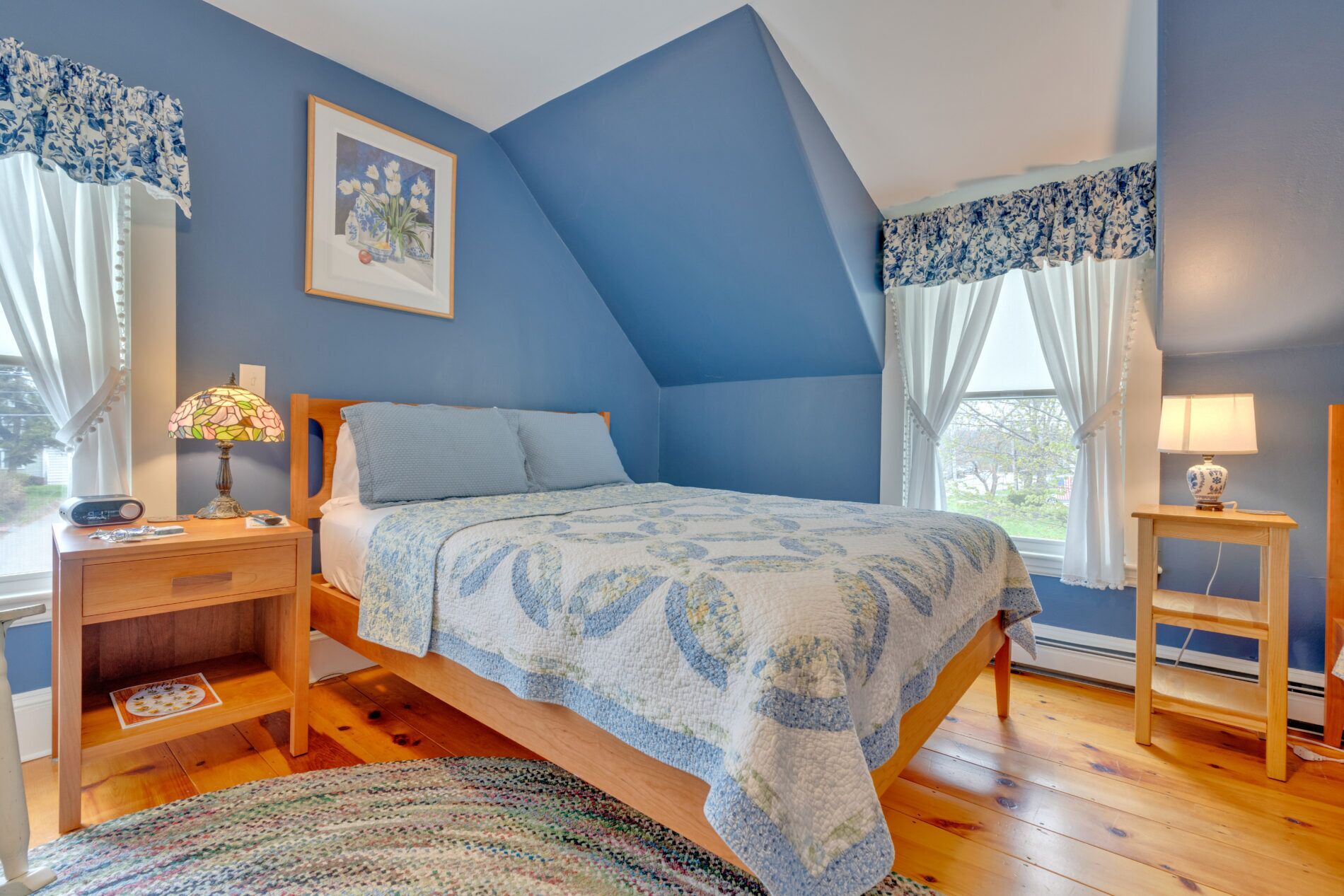Un dormitorio con cama, mesitas de noche, lámpara, un cuadro enmarcado y una ventana con cortinas.