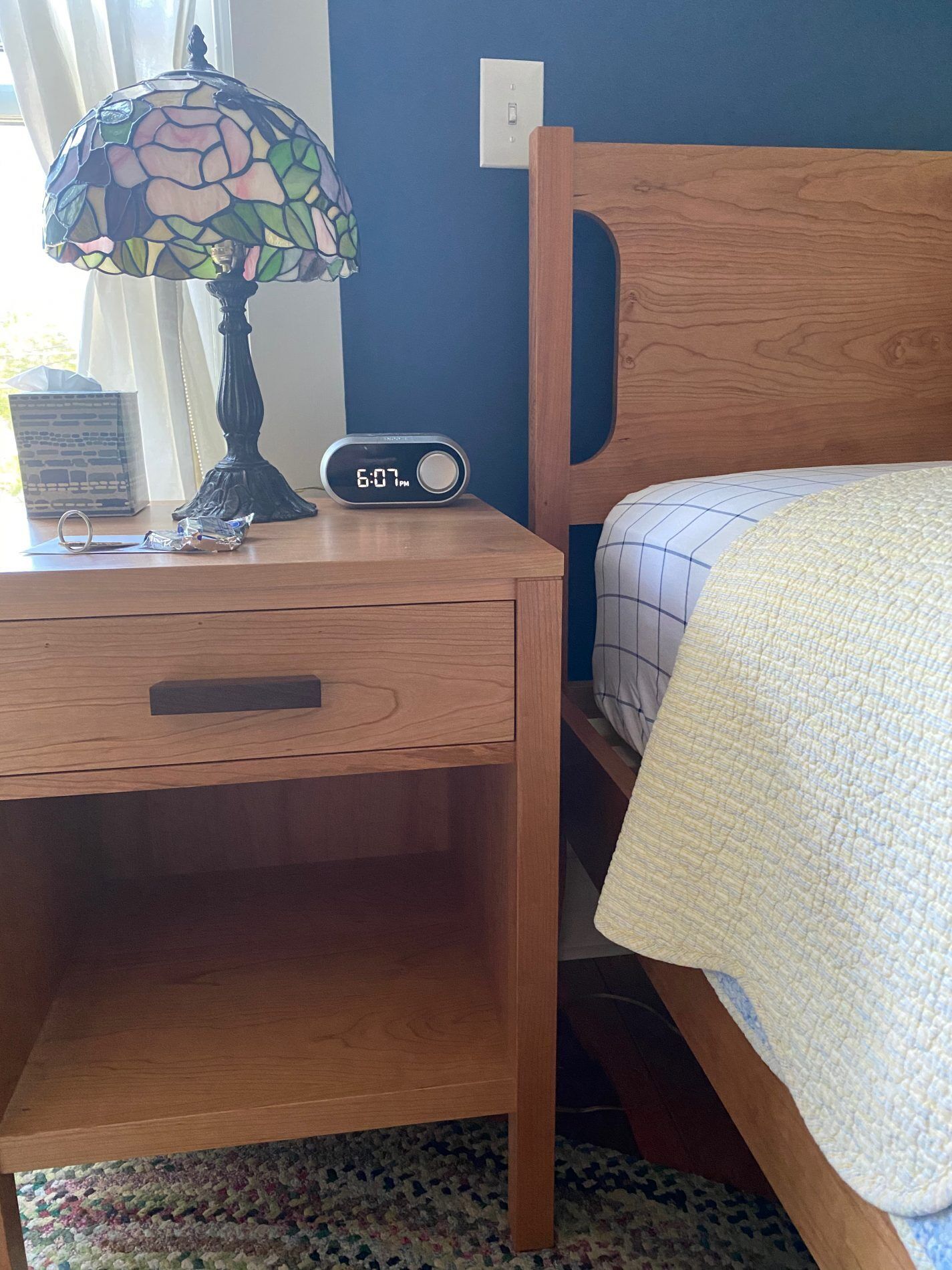 A wooden bedside table with a clock, a lamp, and a bed with a quilt.