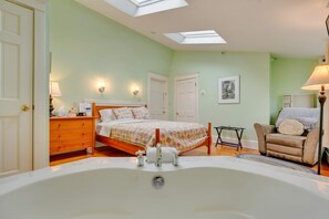 A bedroom with a large bathtub, a bed with floral bedding, a chair, and a desk with a lamp. - The James Place Inn B&B (Freeport)