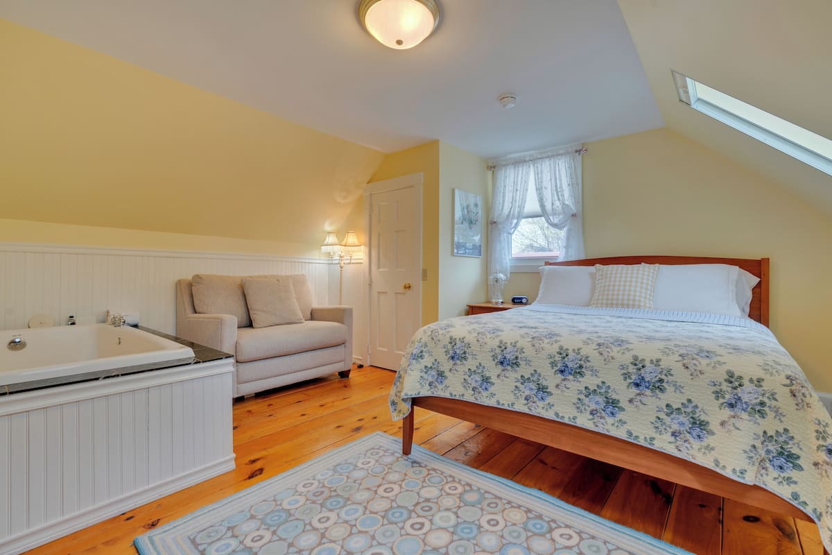 A bedroom with a wooden bed, a white bathtub, a beige armchair, and a small table with a lamp.