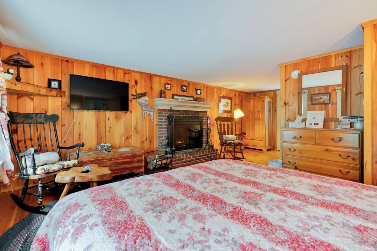 A wooden-paneled room with a fireplace, a bed with a floral bedspread, a rocking chair, a wooden dresser, and a television mounted on the wall.