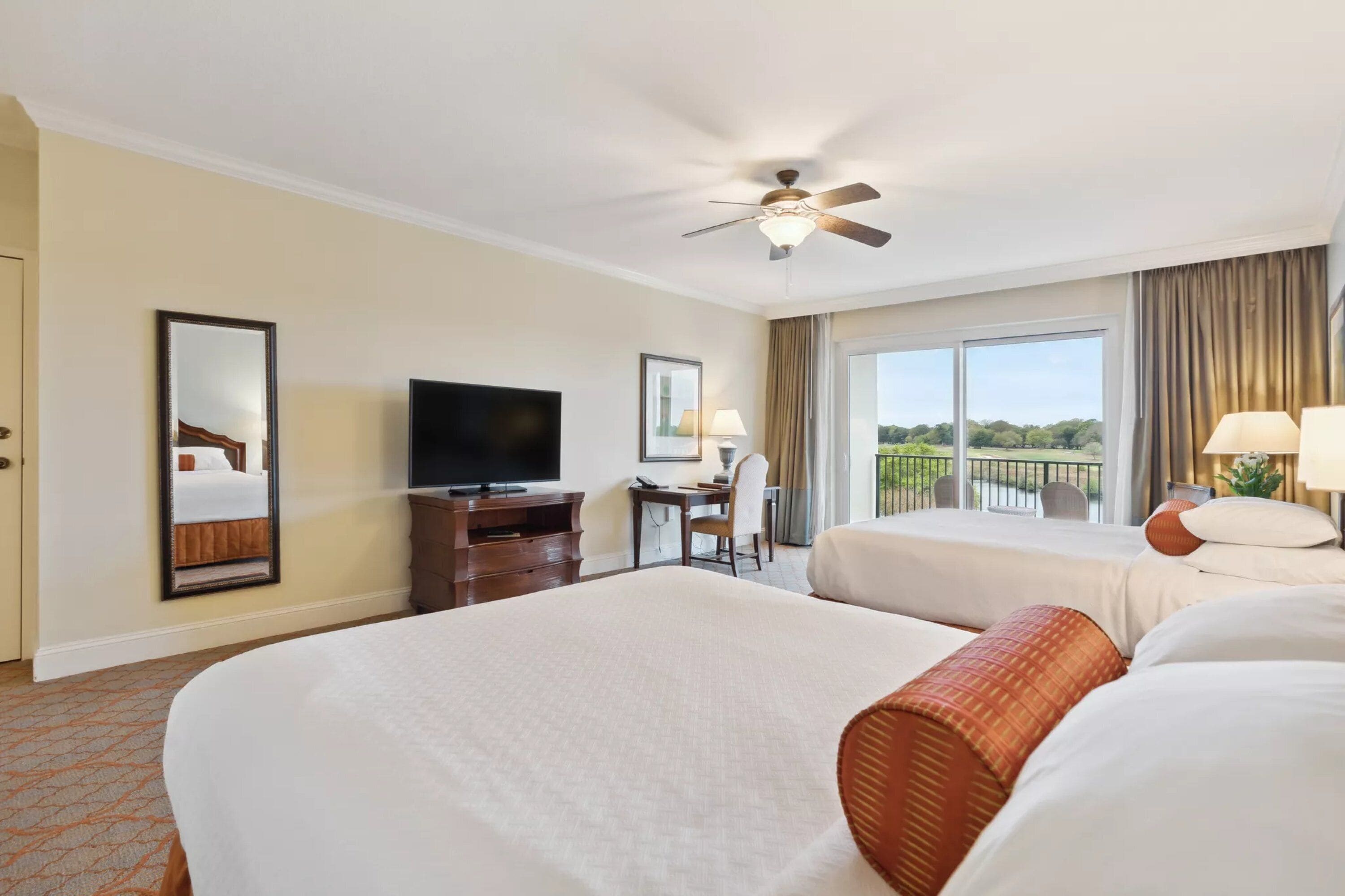 A hotel room with two beds, a desk, a television, and a balcony with a view.
