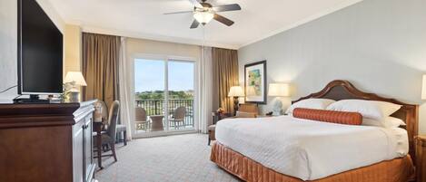 A hotel room with a large bed, a ceiling fan, a TV, a desk, and a balcony with chairs.