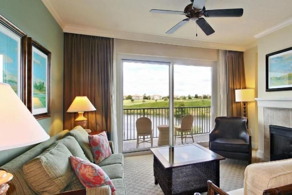 A hotel room with a balcony, a sofa, a chair, a table, a fireplace, and a ceiling fan.