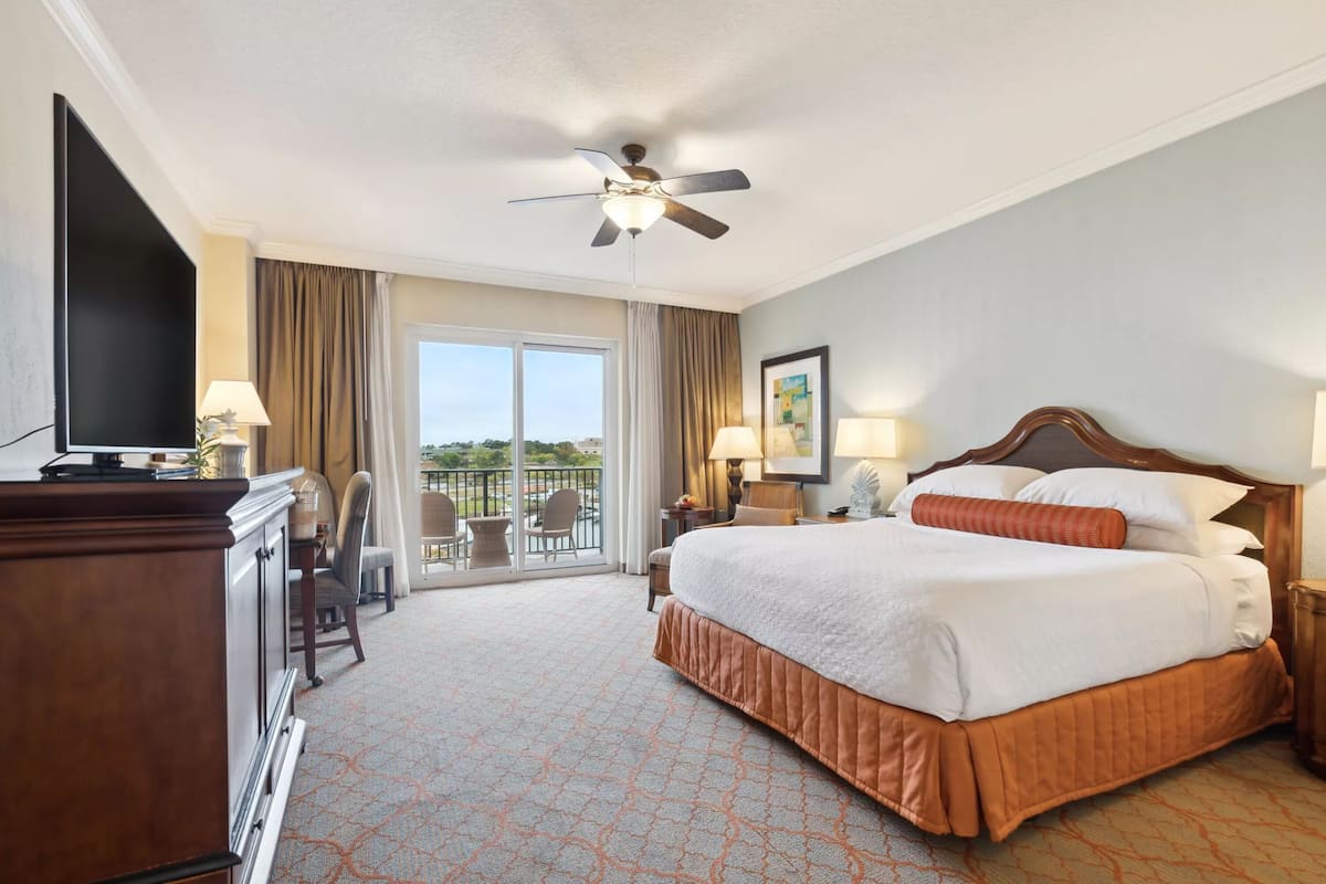 A hotel room with a large bed, a ceiling fan, a TV, a desk, and a balcony with chairs.