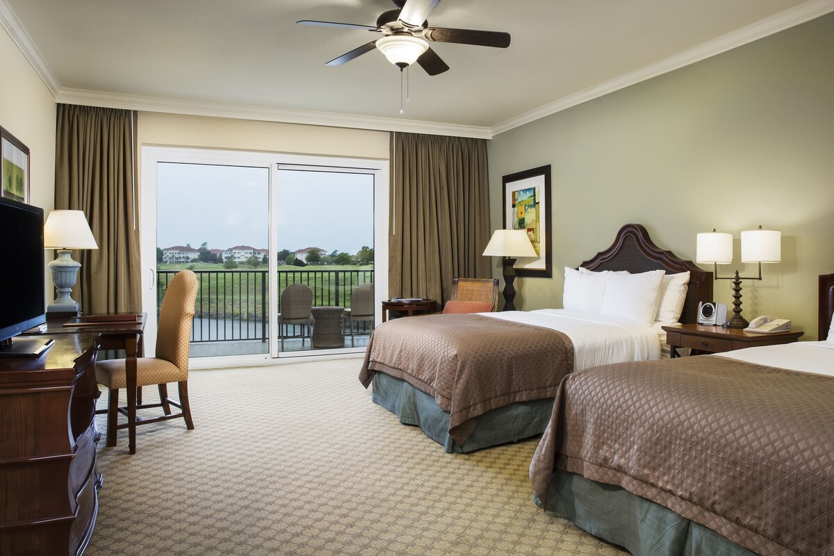 A hotel room with two beds, a desk with a chair, a television, a ceiling fan, and a view of a golf course.