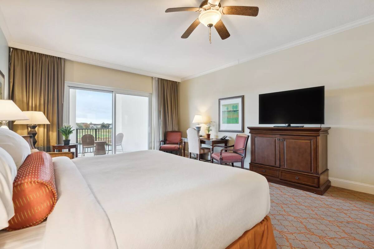 A hotel room with a large bed, a ceiling fan, a TV, a desk with a chair, and a view of the outdoors.
