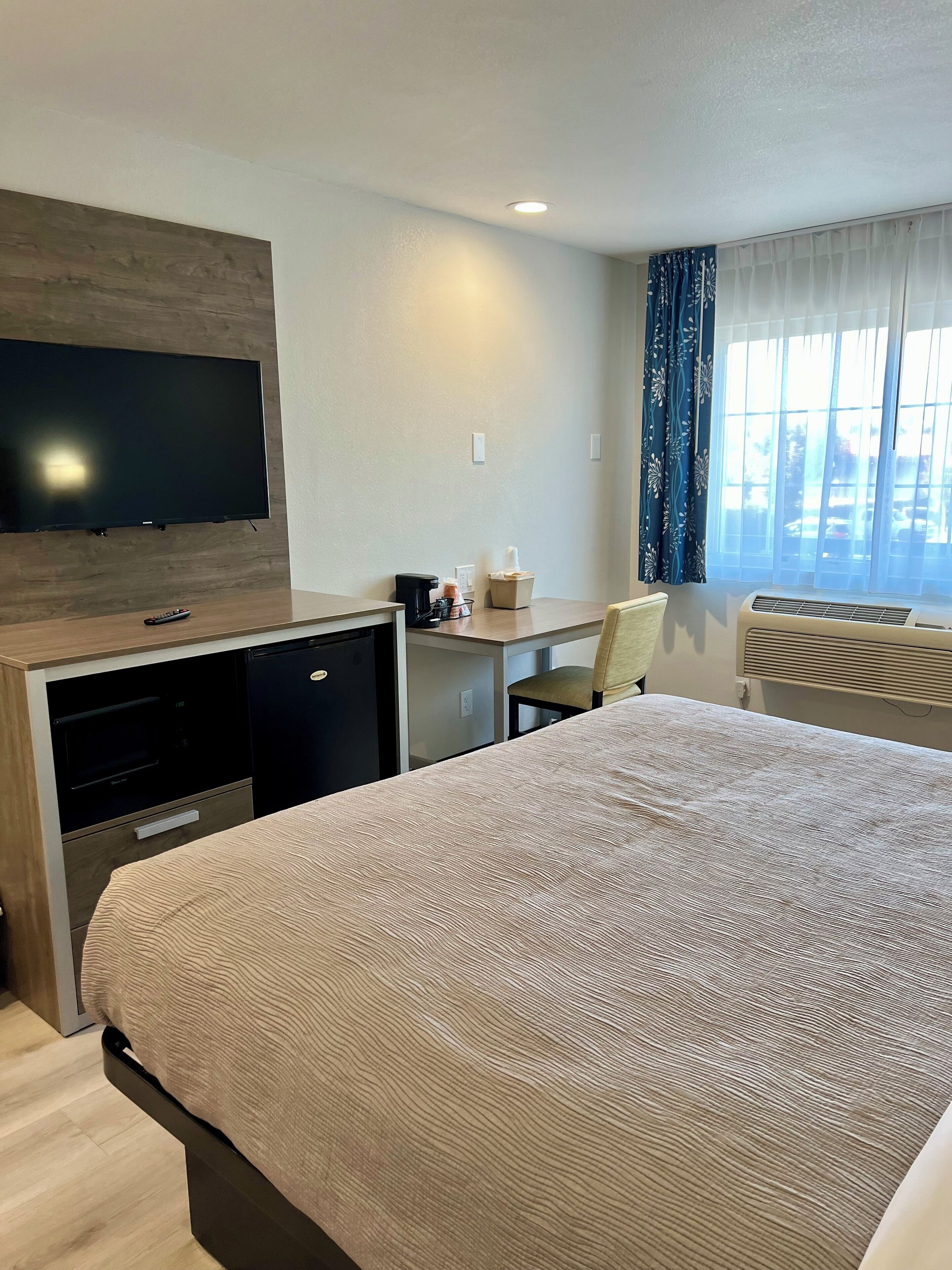 A hotel room with a bed, a desk, a TV mounted on the wall, a coffee maker, and a window with curtains.