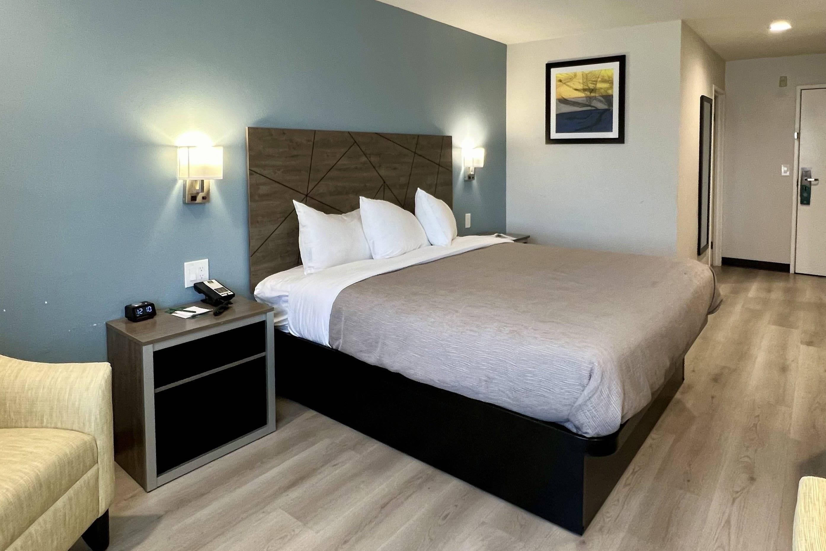A hotel room with a bed, bedside tables, a nightstand, a chair, and a painting on the wall.