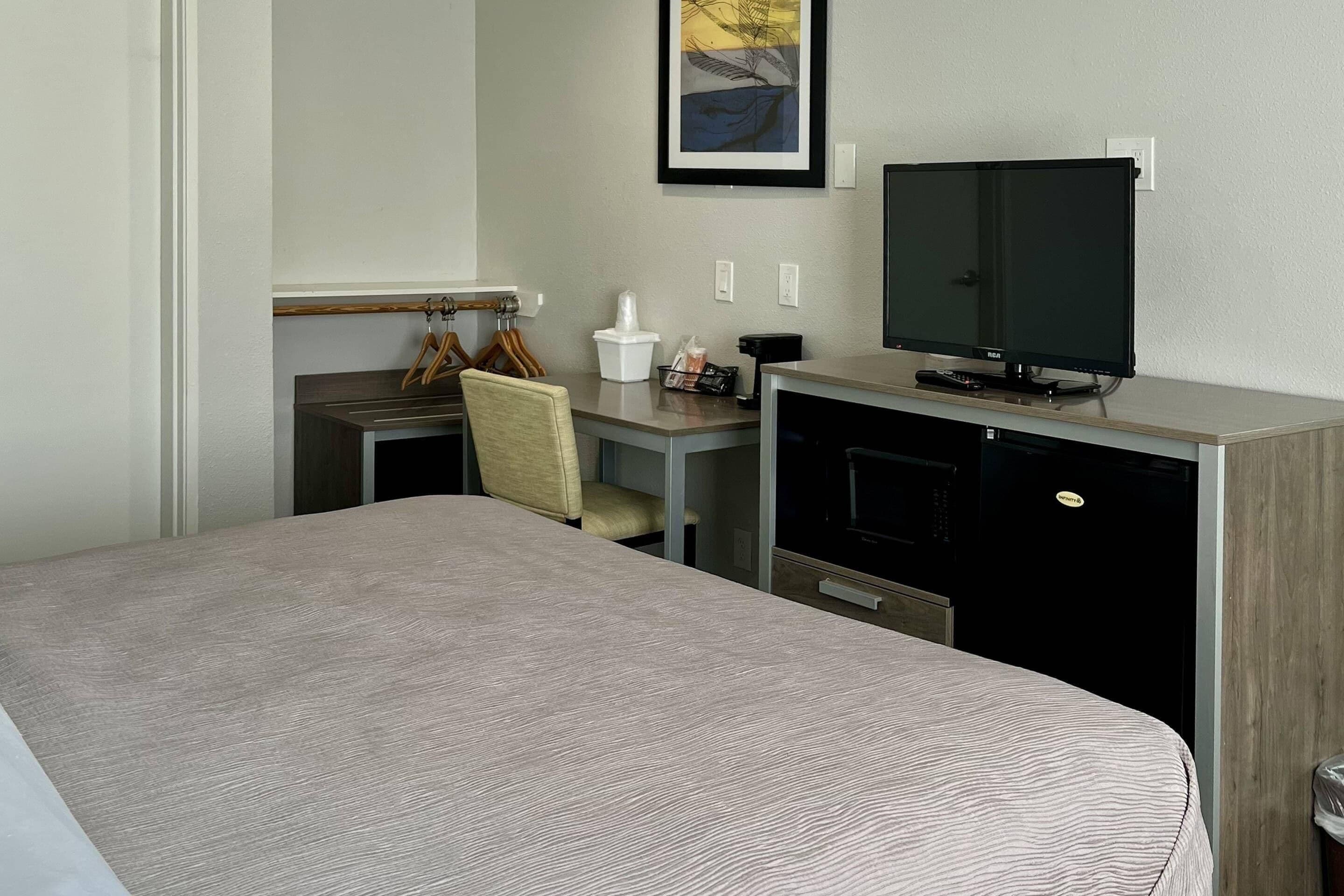A hotel room with a bed, a desk with a television, a chair, and a small kitchenette area.